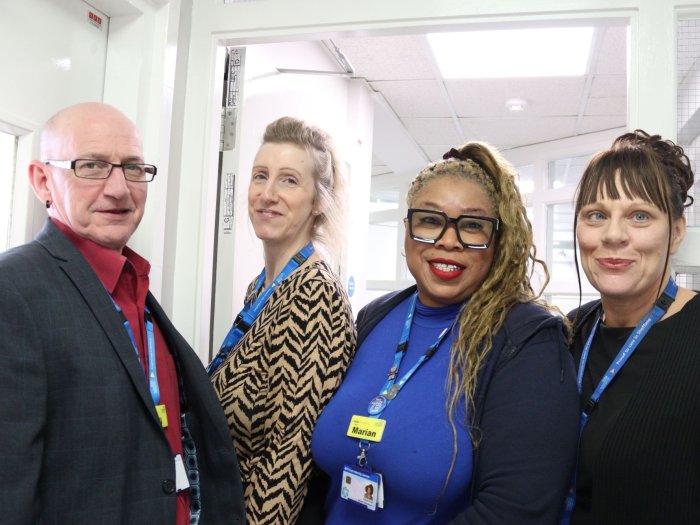 Four people in semi-formal wear stand together side by side in a corridor smiling at the camera, wearing NHS lanyards